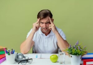 Stressed man with books, apple, and plant on desk.